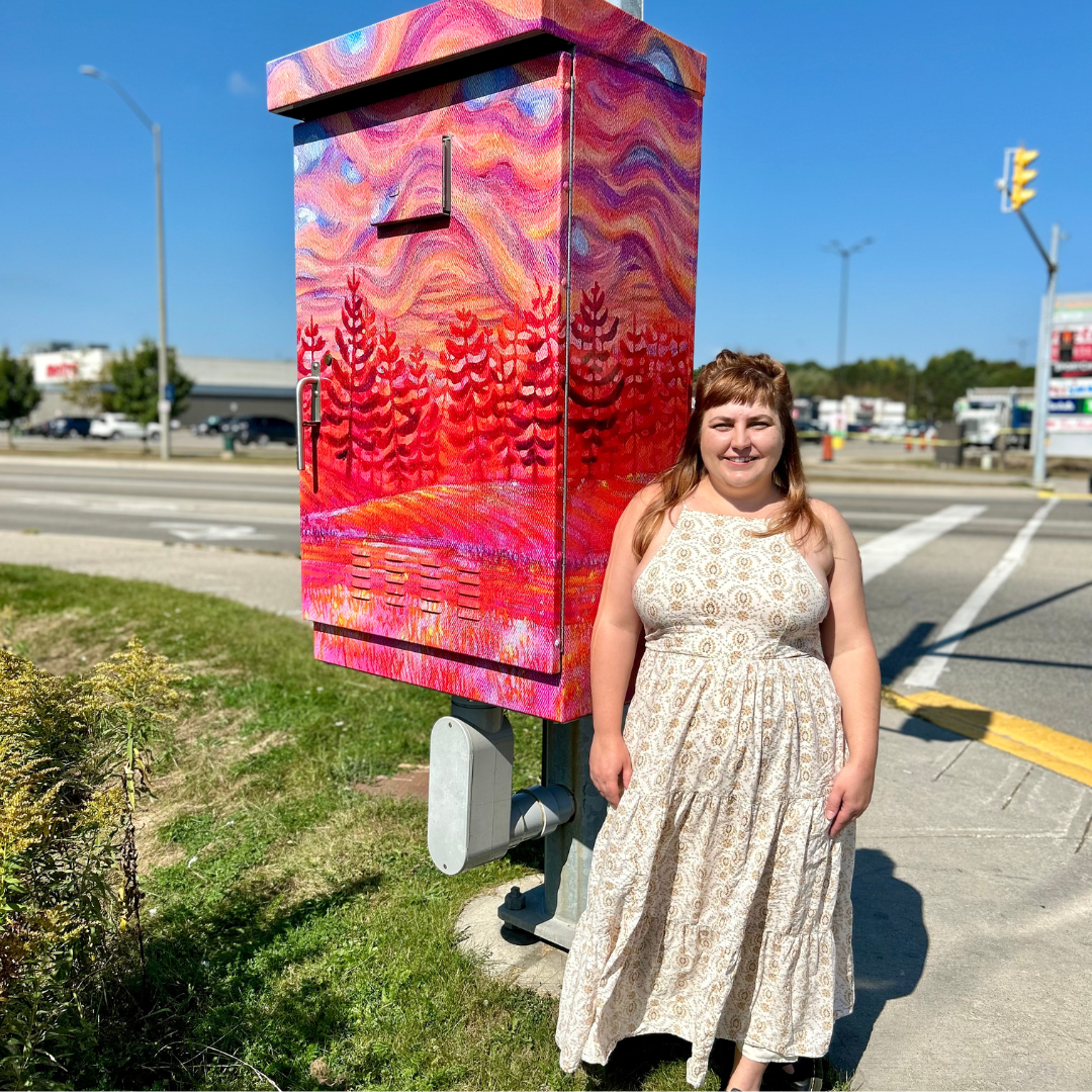 A women stands in front of a utility box wrapped in art depicting a trees and a landscape at sunset.